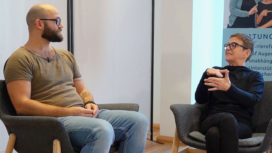 Interview mit Christine Christine Linnartz und ein Gebärdensprachdolmetscher nebeneinander sitzend. Sie haben einander den Blick zugewandt.
