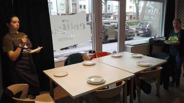 Das Bild zeigt einen Innenraum. In der Mitte des Bildes befinden sich zwei zusammengeschobene Tische mit 6 Stühlen. Der Tisch ist mit 6 Tellern gedeckt. Auf der linken Seite hält eine junge Frau mit einer schwarzen Schürze einen Teller fest. Auf der rechten Seite hält eine junge Frau zwei Teller fest.