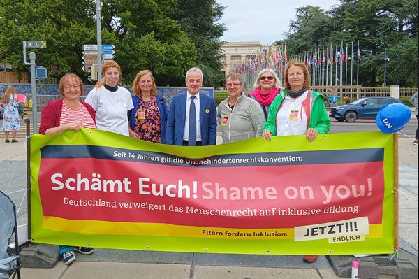 Sieben Personen stehen hinter einem Transparent. In der Mitte steht ein Herr mit dunkelblauem Anzug und Krawatte. Auf dem Transparent steht „Seit 14 Jahren gilt die UN-Behindertenrechtskonvention: Schämt Euch! Shame on you! Deutschland verweigert das Menschenrecht auf inklusive Bildung. Eltern fordern Inklusion. Jetzt! Endlich!!!“