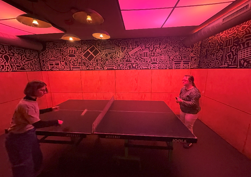 Tischtennis bei Rotlicht Das Bild zeigt einen Raum mit zwei Personen, die sich an einer Tischtennisplatte gegenüber stehen. Die jungen Frauen haben einen Schläger in der Hand und spielen Tischtennis. Der Raum ist rot ausgeleuchtet.