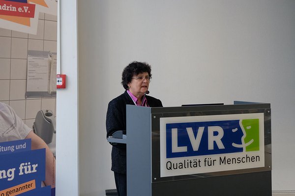 Ein Frau mit dunklen Haaren steht hinter einem Rednerpult und hält einen Vortrag. Auf dem Rednerpult steht LVR.