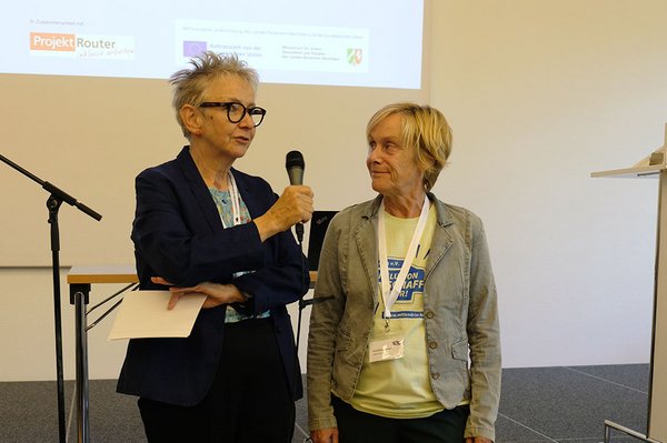 Die Moderatorin Cornelia Benninghoven und Projektleiterin Eva-Maria Thoms stehen vor der Bühne und unterhalten sich. Im Hintergrund ist das Projekt-Logo mit dem Förderlogo von Land NRW und Europäischer Union an die Wand projiziert.