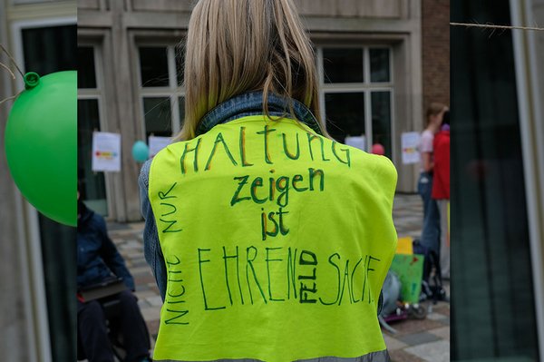Rückenansicht einer Person in einer gelben Warnweste. Darauf steht handschriftlich: „Haltung zeigen ist Ehrensache. Nicht nur." Im Hintergrund findet die Demonstration statt.