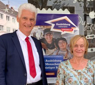 Großer mittelalter Mann mit grauem Haar, Anzug und Krawatte und kleinere mittelalte Frau mit blondem Haar vor dem Fenster eines Ladenlokals, beide Lächeln. Im Fenster hängt ein Plakat mit der Aufschrift "Ausbildung mittendrin"