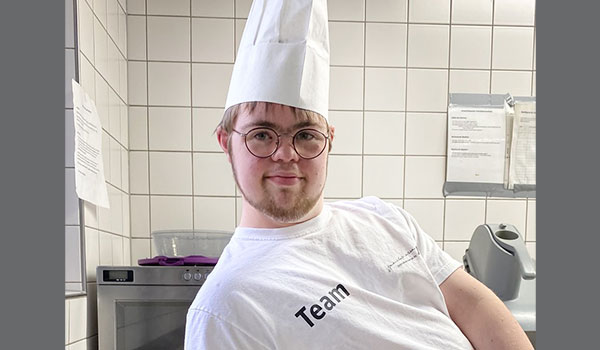 Auszubildener Lucas Ein junger Mann mit Down-Syndrom lächelt in die Kamera. Er hat eine weiße Kochmütze und ein weißes T-Shirt mit dem Schriftzug Team an. Er lehnt an einer Küchenarbeitsplatte und hat eine Kiwi geschnitten.