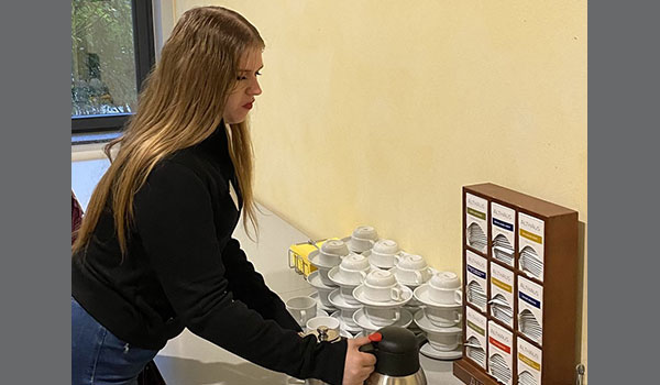 Links im Bild ist eine junge Frau zu sehen, die einen Tisch mit Catering vorbereitet. Sie stellt eine Kanne Kaffee auf den Tisch. Auf diesem Tisch befinden sich außerdem Tassen, Milch und Tee. 