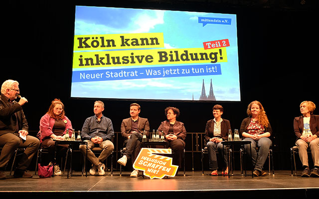 Acht Persoenen sitzen nebeneiandenr aufeine rBühne. Einer spricht in ineMikrofon. Im Hintergrund ist ein Bild projiziert. Auf einem gelben Balken steht in schwarzer Schrift: „Köln kann inklusive Bildung - Teil 2. Neuer Stadtrat – Was jetzt zu tun ist."