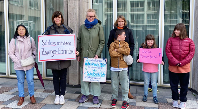 Vier Erwachsene und drei Kinder mit Down-Syndrom stehen vor einem Gebäude. SIe haben 2 Schilder in der Hand. Auf einem steht „Schluss mit dem Zwangs-Elterntaxi“. Auf dem anderen Schild steht „Schafft endlich mehr wohnortnahe  Plätze an inklusiven Schulen oder einen Schulbusverkehr ins Gemeinsame Lernen für Schüler:innen mit Behinderung!"