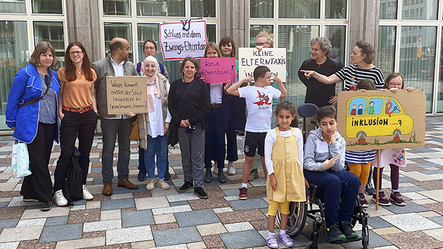 Elf Erwachsene und fünf Kinder stehen vor einem Gebäude. Ein Kind sitzt im Rollstuhl. Drei Kindern haben Down-Syndrom. Sie halten Schilder mit folgenden Aufschriften hoch: „Schluss mit dem Zwangs-Elterntaxi!", Kiene Elterntaxis in die Inklusion, „Wann kommt endlich der Schulbusverkehr ins Gemeinsame Lernen?"