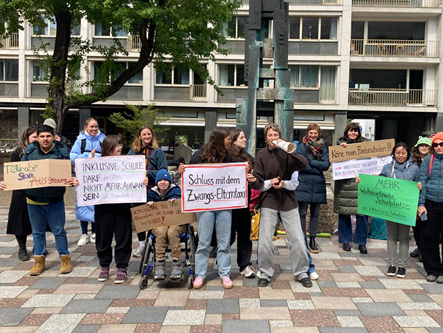 Etwa 20 Jugendliche und Erwachsene mit und ohne Behinderung stehen auf einem Platz und halten Schilder mit folgenden Aufschriften: Teilhabe statt Ausgrenzung, Inklusive Schule darf nicht mehr Aufwand sein, Gerechtigkeit für alle Menschen ist wichtig, Schluss mit dem Zwangs-Elterntaxi!, Keine neue Förderschulen, mehr wohnortnahe Plätze an inklusiven Schulen.